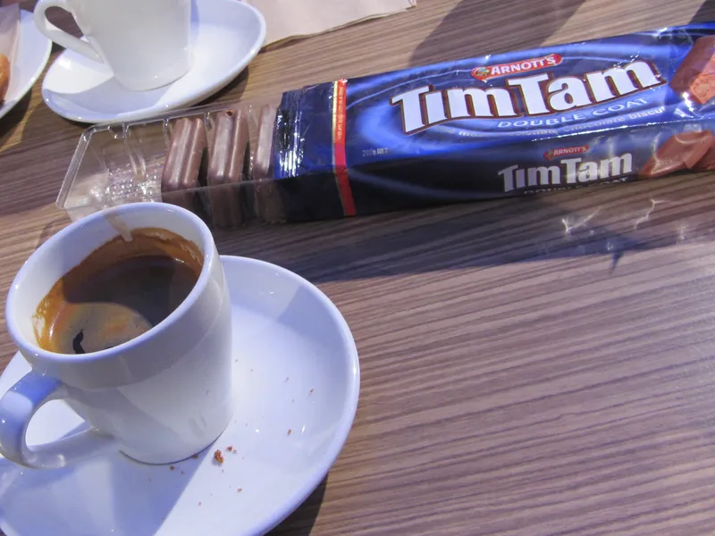 A small white cup of black coffee on a saucer in front of an open blue packet of Tim Tam Double Coat biscuits on a wooden cafe table