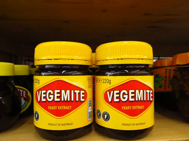 Two 220 g (7.7 oz) jars of Vegemite with yellow lids and red diamond labels on a wooden store shelf, a jar of fig spread beside them