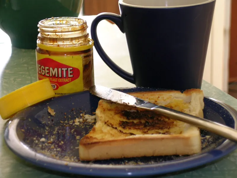 A slice of buttered toast with a thin scrape of Vegemite on a blue plate, a knife resting on it, an open Vegemite jar and a dark blue coffee mug behind