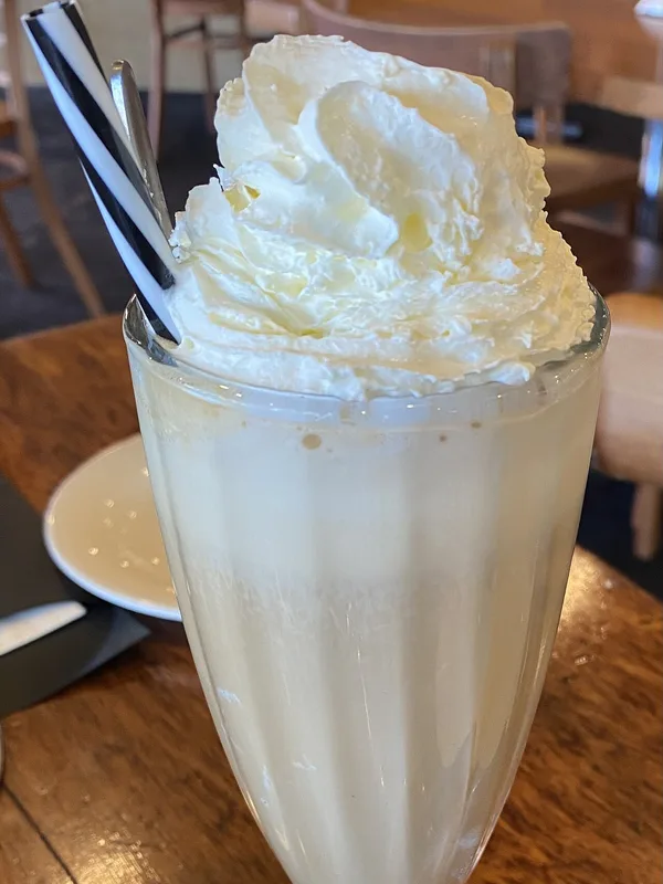 A tall glass of iced coffee with ice cream, topped with whipped cream and a striped straw, on a wooden café table