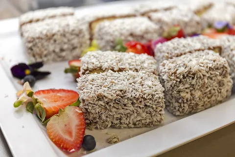Lamingtons, squares of sponge coated in chocolate and coconut, on a white platter with sliced strawberries
