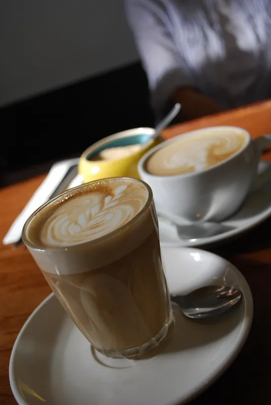 A caffè latte in a tall glass in front, with a flat white in a white ceramic cup behind it, on a café table in Melbourne