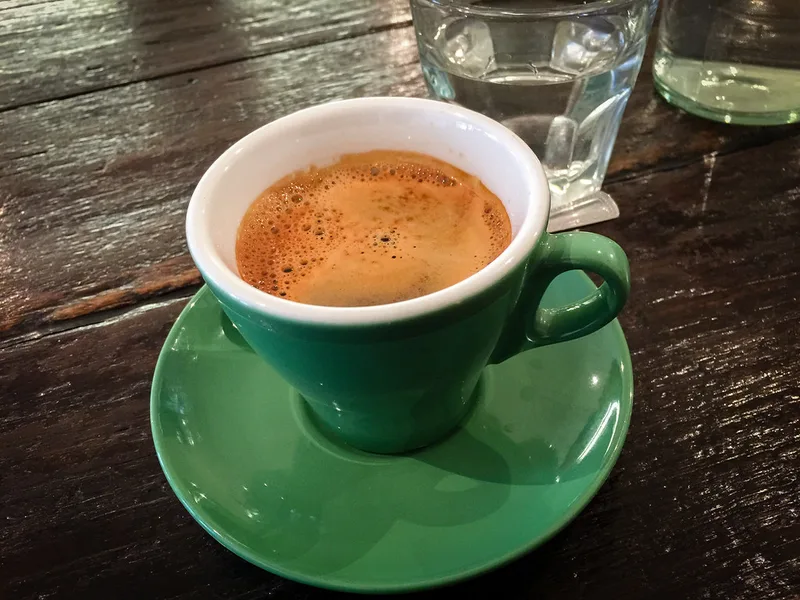 A long black with thick golden crema in a green cup and saucer on a dark wooden café table, with a glass of water behind