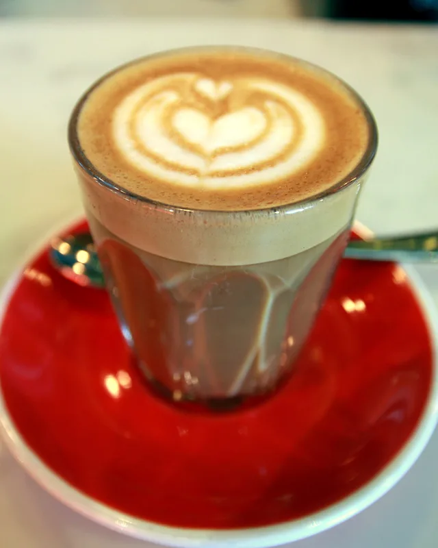 A piccolo latte with heart latte art in a small glass tumbler on a red saucer