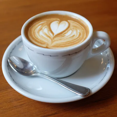 A cappuccino with heart latte art in a white ceramic cup on a saucer with a spoon