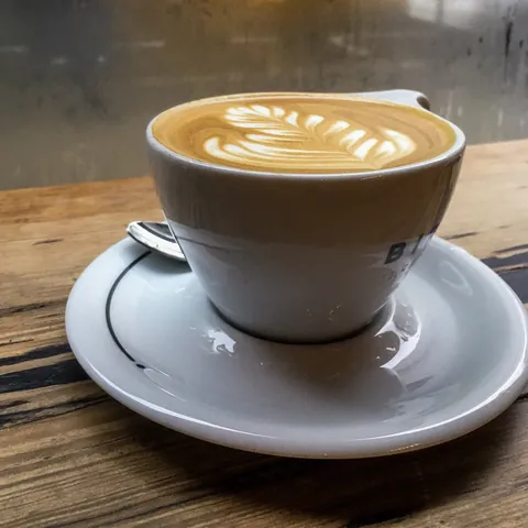 A latte with leaf latte art in a white ceramic cup on a saucer, on a wooden café bench